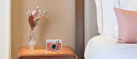 A neatly arranged hotel room with a bed, a bedside table with a camera, and a vase with dried flowers.