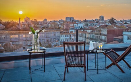 A terrace with a view of a cityscape at sunset, featuring two chairs and a table with a vase of flowers.