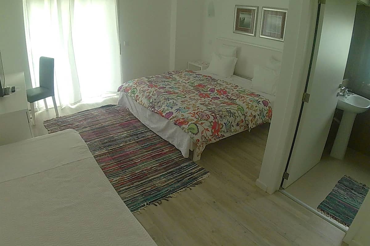 A bedroom with a bed, a chair, a desk, a window with curtains, and a bathroom with a sink.