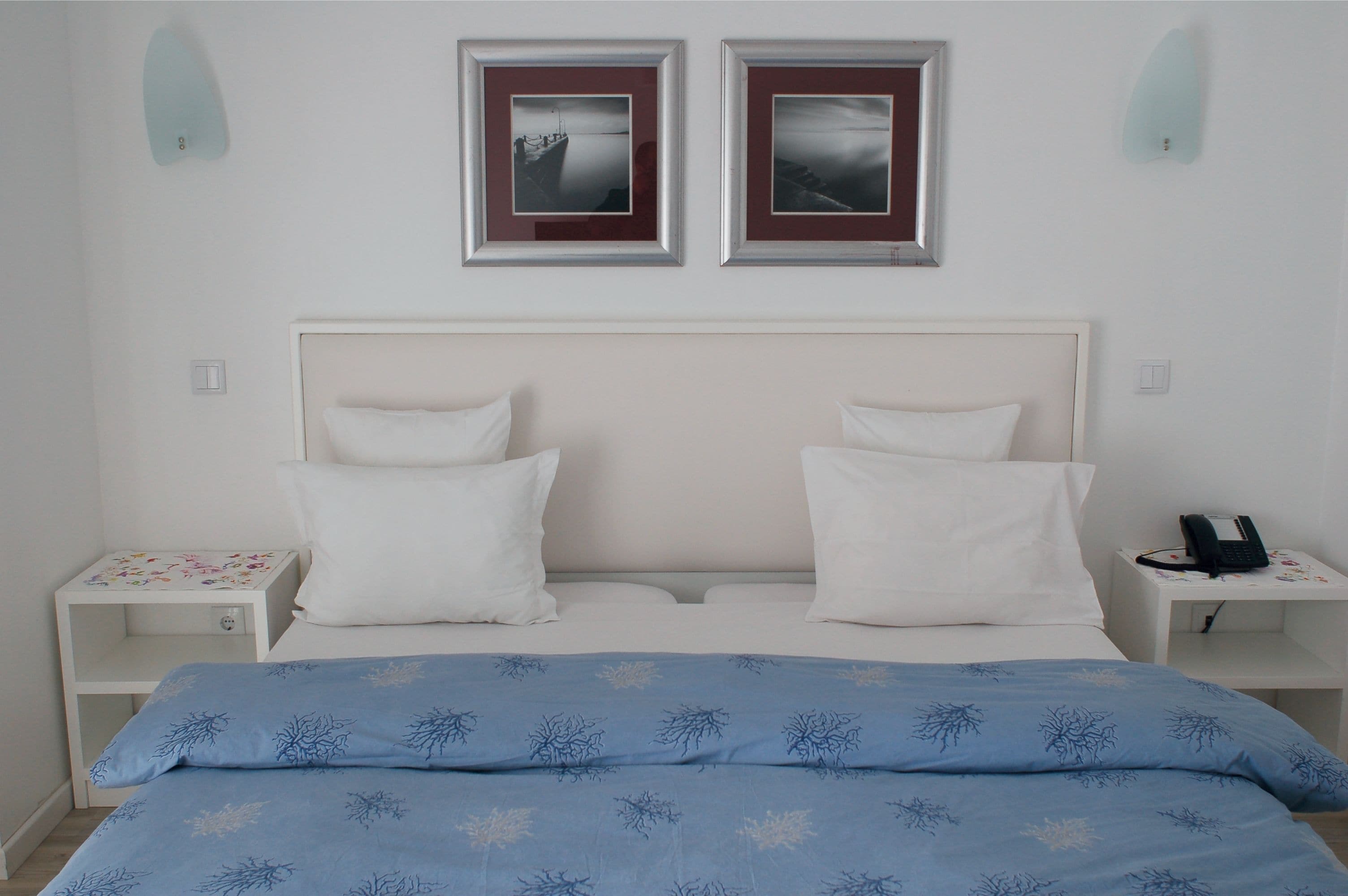 A neatly made bed with white and blue bedding, two bedside tables with lamps, and two framed pictures on the wall.