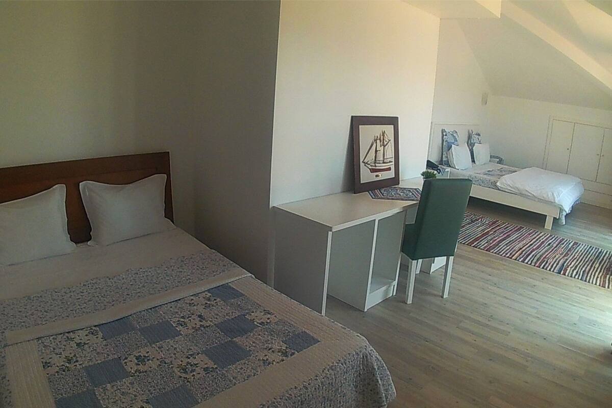 A bedroom with a bed, a desk, and a chair. There is a window with a view of the outside.
