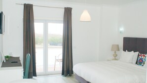 A bedroom with a large bed, a bedside table with a lamp, a chair, and a view through a glass door. - Hotel Baleal Spot (Peniche)