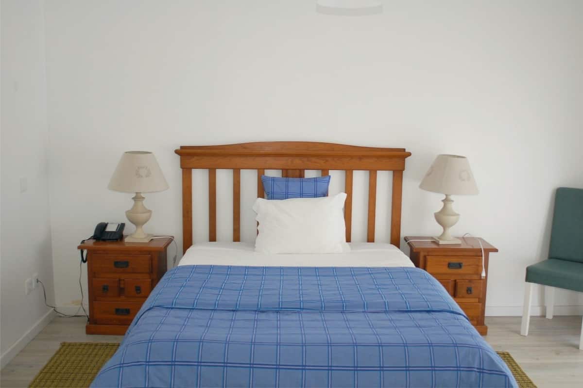 A bedroom with a wooden headboard, a blue checkered bedspread, two bedside tables with lamps, a green chair, and a phone on one of the tables.