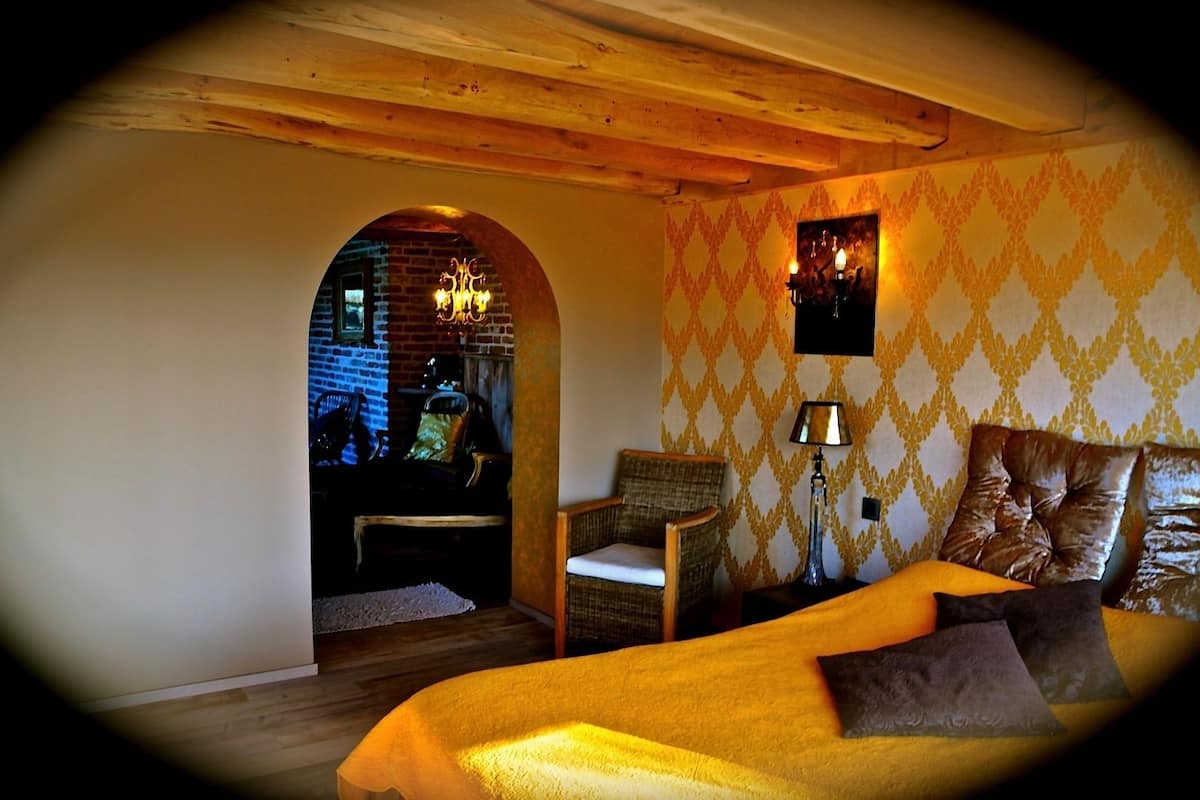 Quarto aconchegante com uma cama, uma luminária e uma cadeira. Apresenta uma parede de tijolos e teto de madeira.