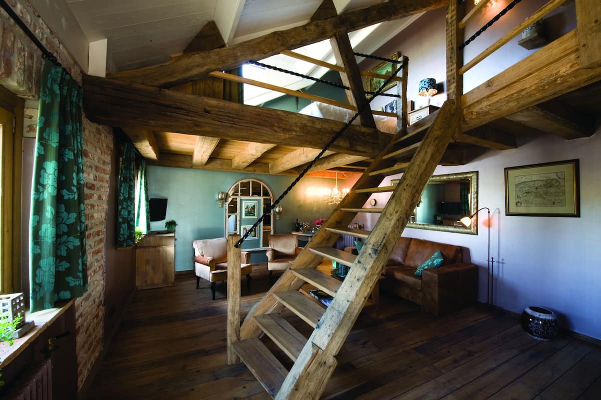 Interior rústico com vigas de madeira aparente, escada de madeira e um espaço de estar aconchegante com lareira.