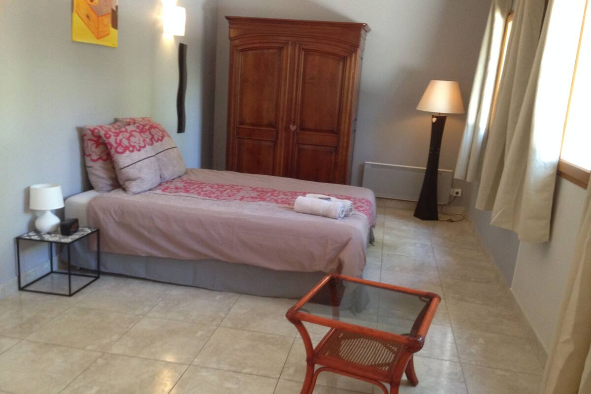 A bedroom with a bed, a nightstand, a wooden wardrobe, a glass table, and a lamp.