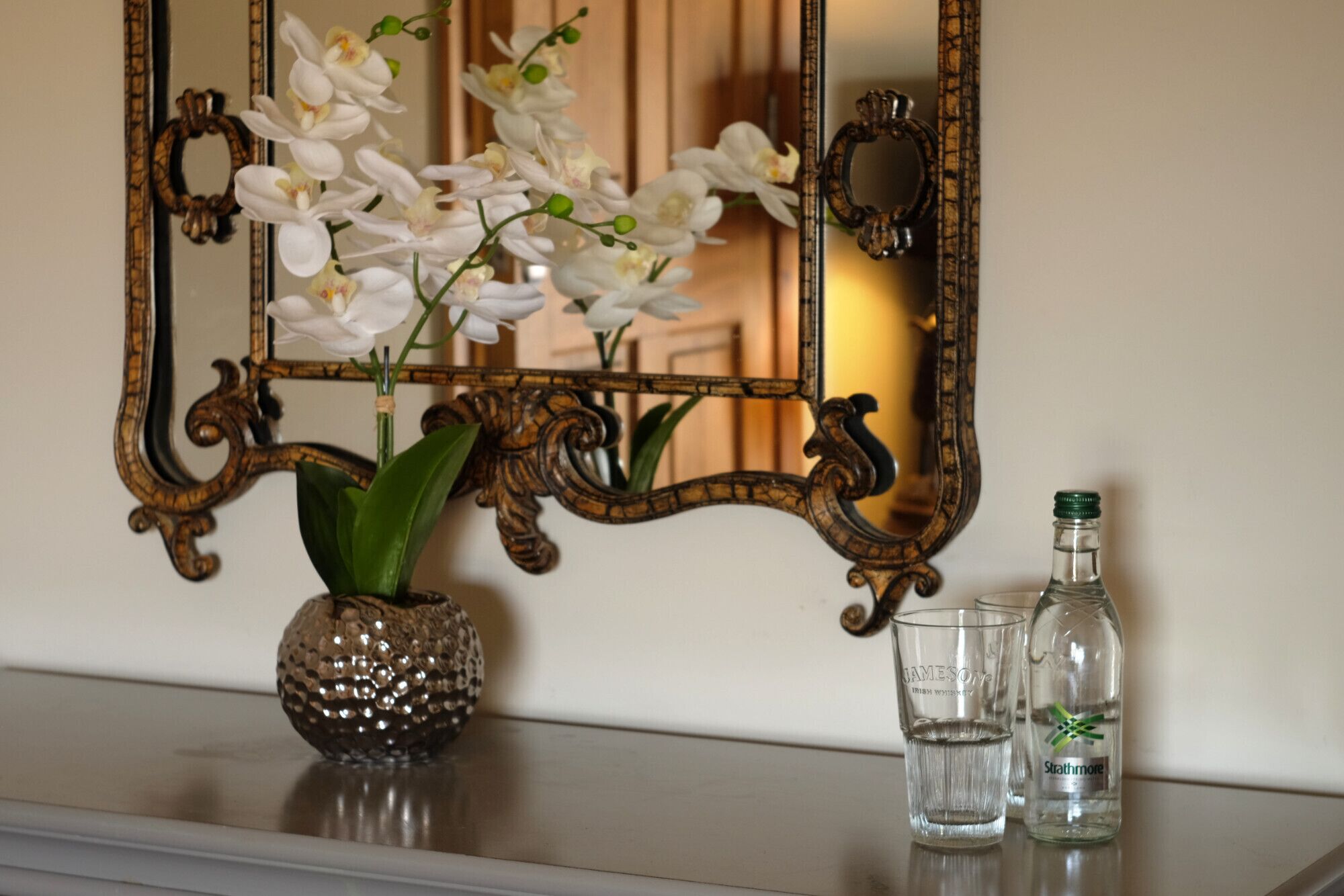 A mirror with ornate wooden frame, a vase with white orchids, and two glasses with a bottle of whiskey.