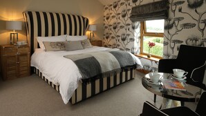 A bedroom with a large bed, bedside tables, a window with a view, and a small table with a chair. - The Queens at Horton (Telford)