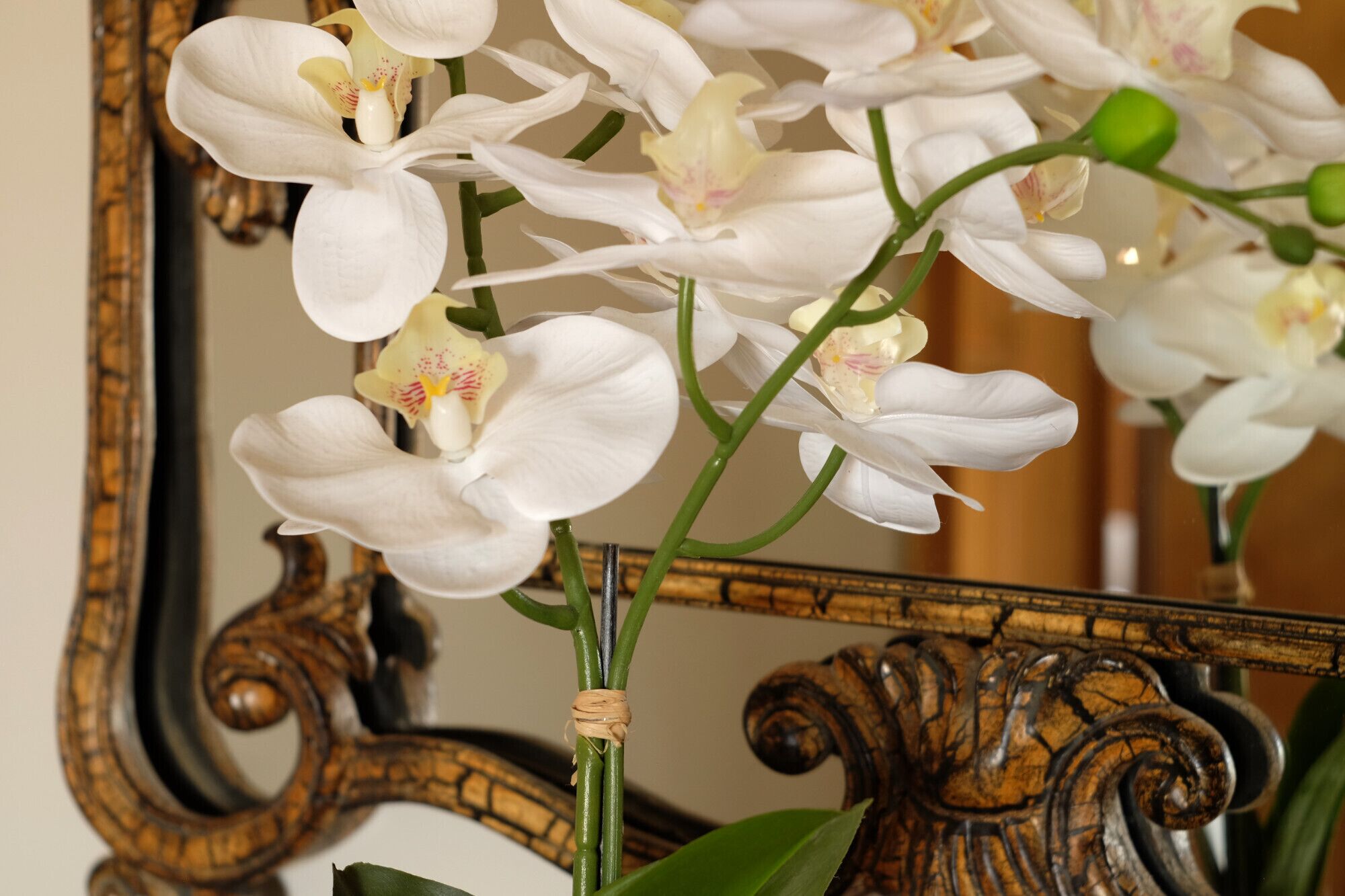 Een close-up van witte orchideeën met gele centrum, vastgebonden aan een groene stengel, voor een houten spiegel met gedetailleerde snijwerken.