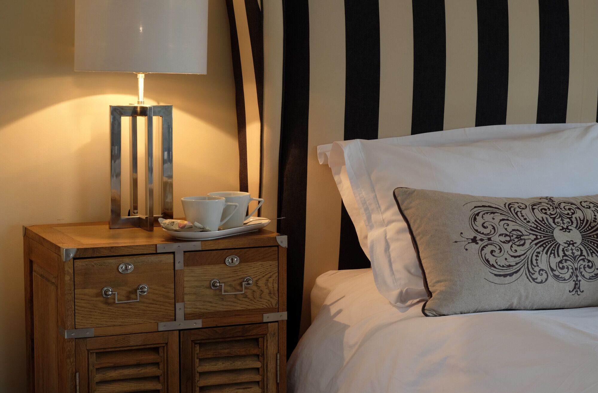 A neatly made bed with a decorative pillow, a bedside table with a lamp, and a tray with cups and a teapot.