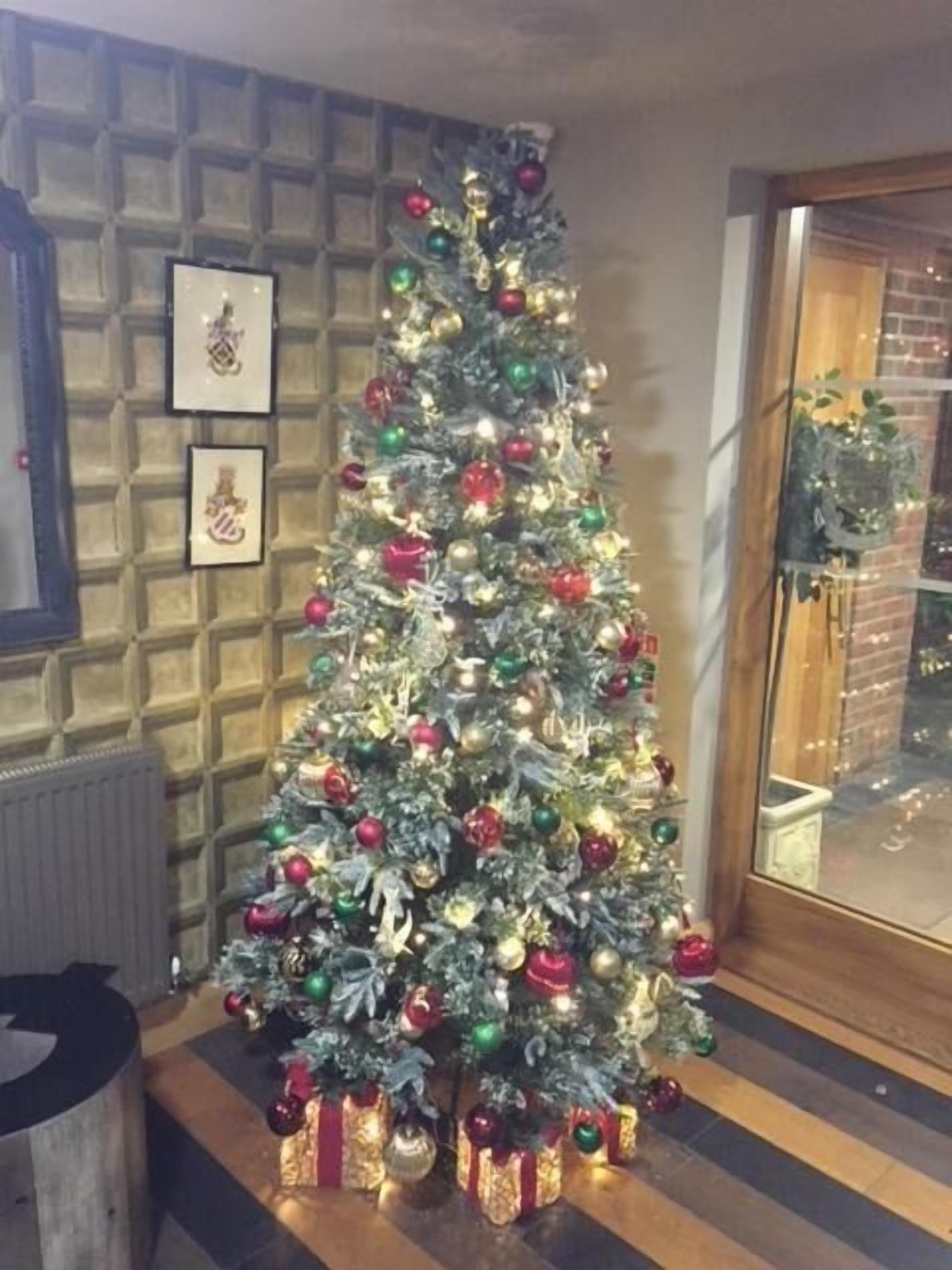 A Christmas tree with ornaments and lights, positioned in a corner with a brick wall and framed pictures.