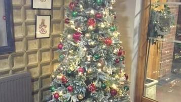 A Christmas tree with ornaments and lights, positioned in a corner with a brick wall and framed pictures.