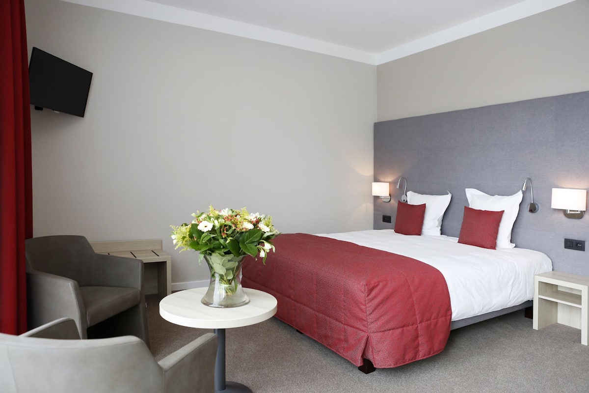 A hotel room with a bed, a desk, a chair, a small table with a vase of flowers, and a TV mounted on the wall.