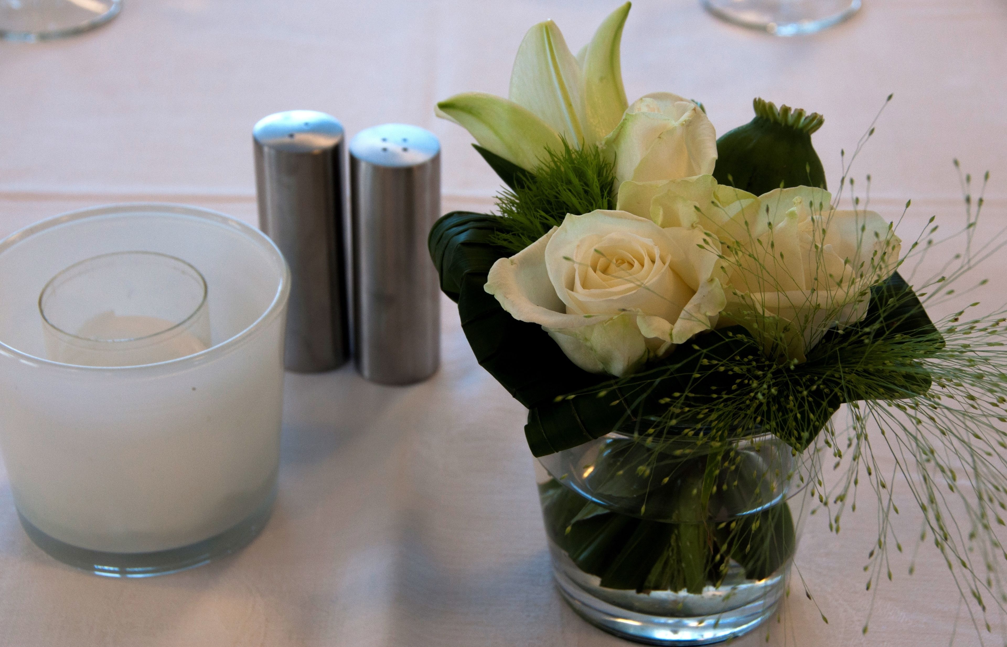 Una mesa dispuesta con una vela en vidrio transparente, un florero de vidrio con flores blancas y dos saleros y pimenteros.