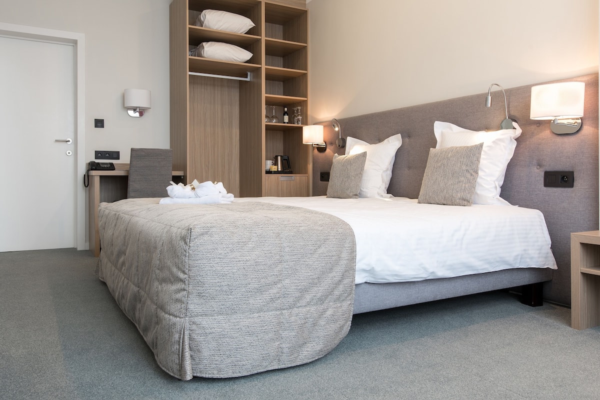 A neatly arranged hotel room with a large bed, a wardrobe, a bedside table, and a wall-mounted lamp.