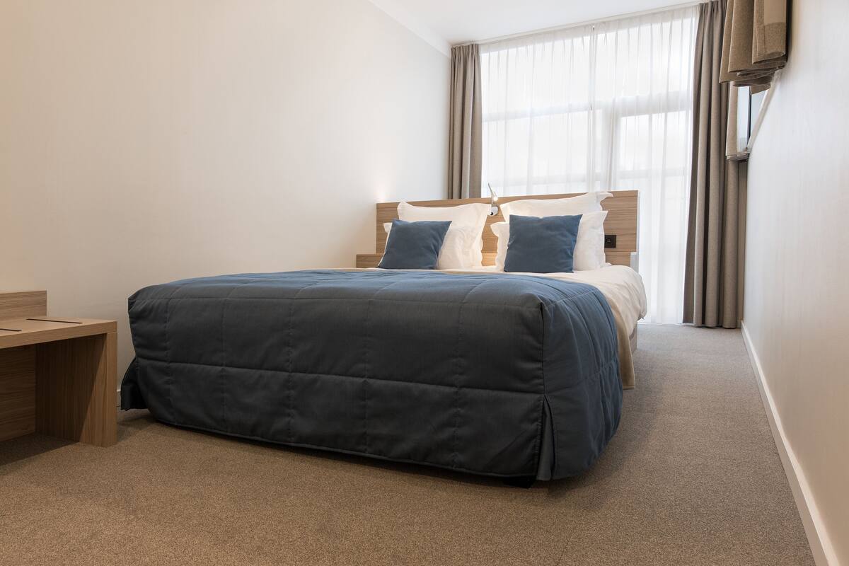 A hotel room with a bed, a wooden nightstand, and a window with curtains.