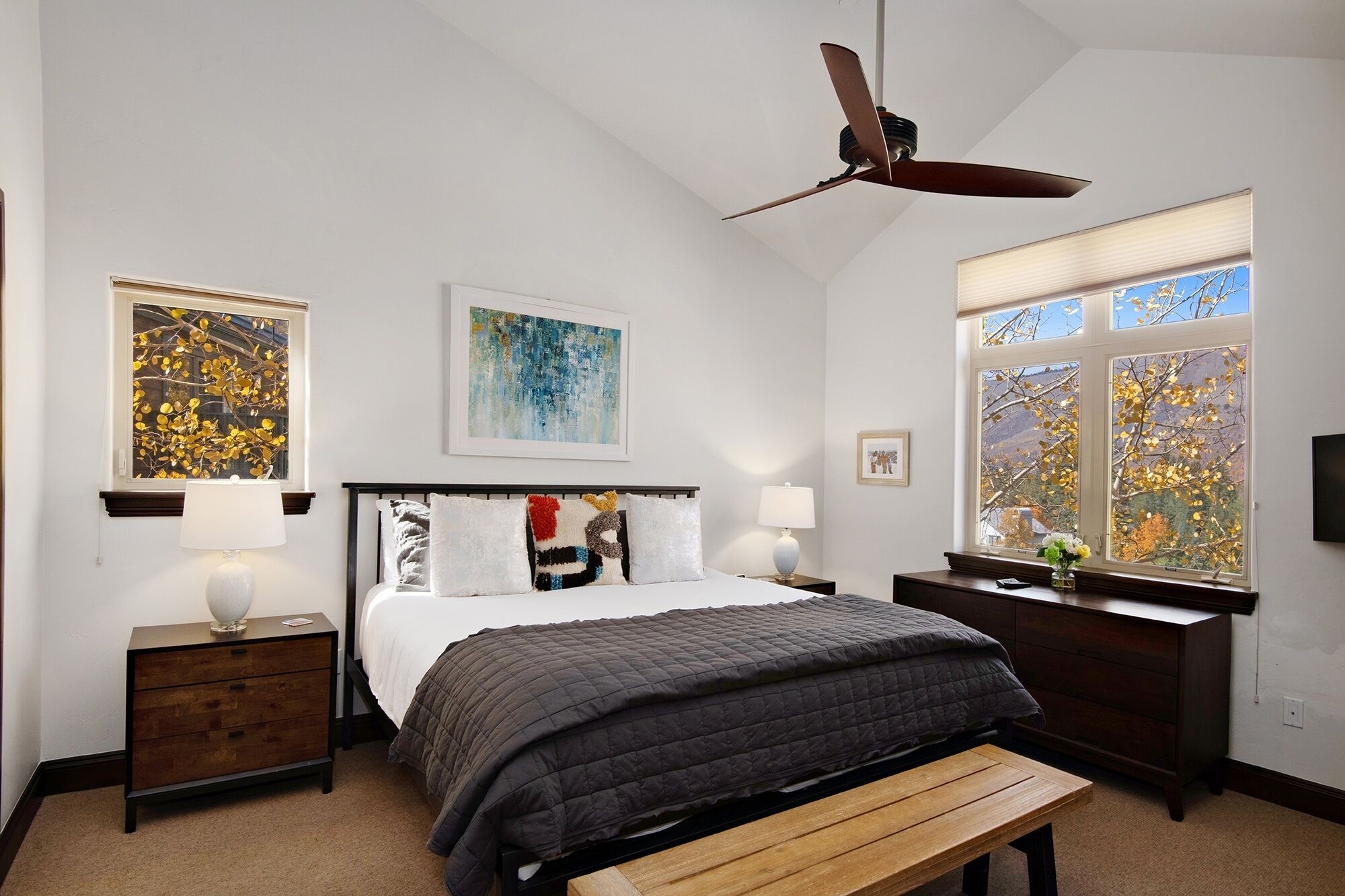 A bedroom with a bed, a wooden bench, a nightstand, a desk, a ceiling fan, and a large window with autumn leaves.