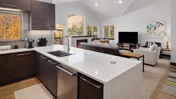 A modern kitchen with a large island, dark wood cabinets, and a white countertop. There is a fireplace, a television, and a seating area with a couch and a coffee table. - Timberfalls 1905 3 Bedroom Condo by RedAwning (Vail)