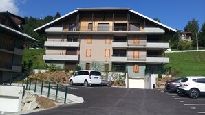 A multi-story building with balconies, a parking area, and a green landscape.