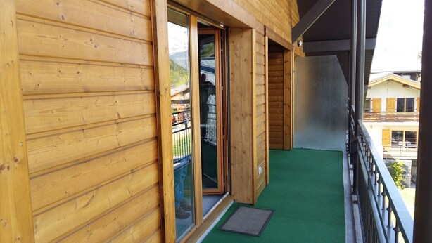 A wooden cabin with a balcony, a sliding glass door, and a green mat at the entrance.