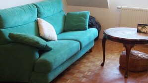 Un canapé turquoise agrémenté de coussins, une table ronde en bois surmontée d’un plateau en verre, et un radiateur en arrière-plan.