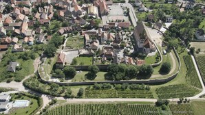 Une vue aérienne d’une ville historique, avec ses bâtiments très rapprochés les uns des autres, une église centrale et des espaces verts environnants.