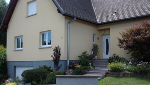 Une maison à deux étages, avec une façade de couleur beige, une partie inférieure grise et un toit recouvert de tuiles. Au premier étage, on aperçoit deux fenêtres, une porte avec un petit porche et un drapeau à l’extérieur.