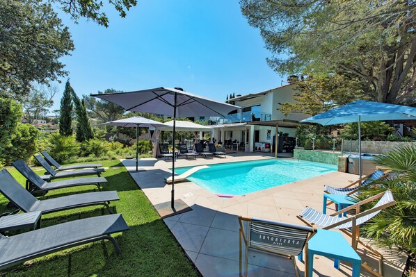 Zona de piscina con sillones reclinables y sombrillas, una casa moderna al fondo y un cielo azul despejado.