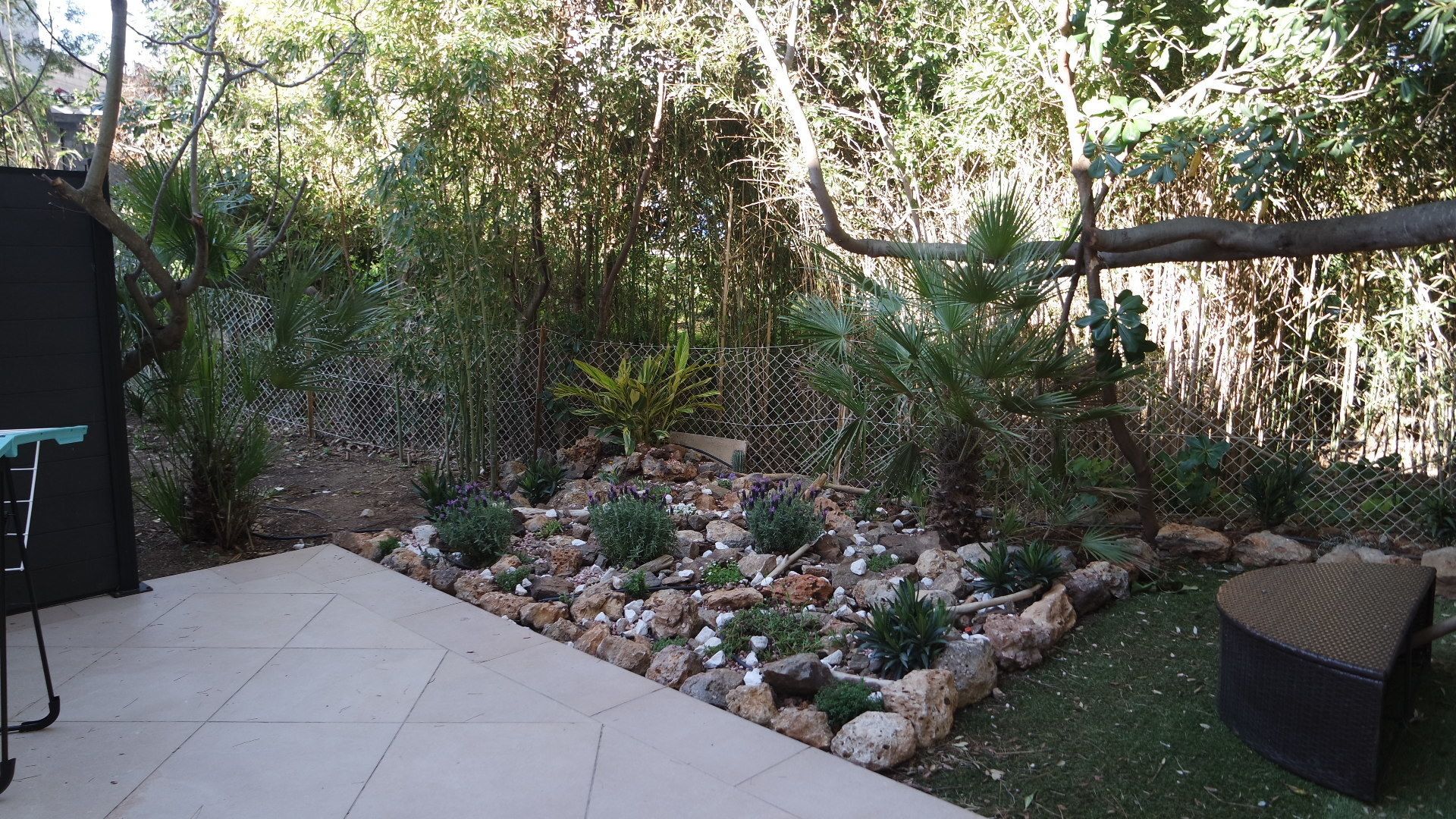 Un patio con suelo de baldosas, una zona de estar con una silla tejida, y un jardín con diversas plantas y rocas.