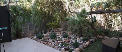 A patio area with a tiled floor, a seating area with a woven chair, and a garden with various plants and rocks.