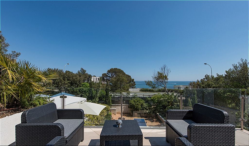 An outdoor seating area with wicker furniture and a white umbrella, overlooking a coastal view with trees and a clear sky.