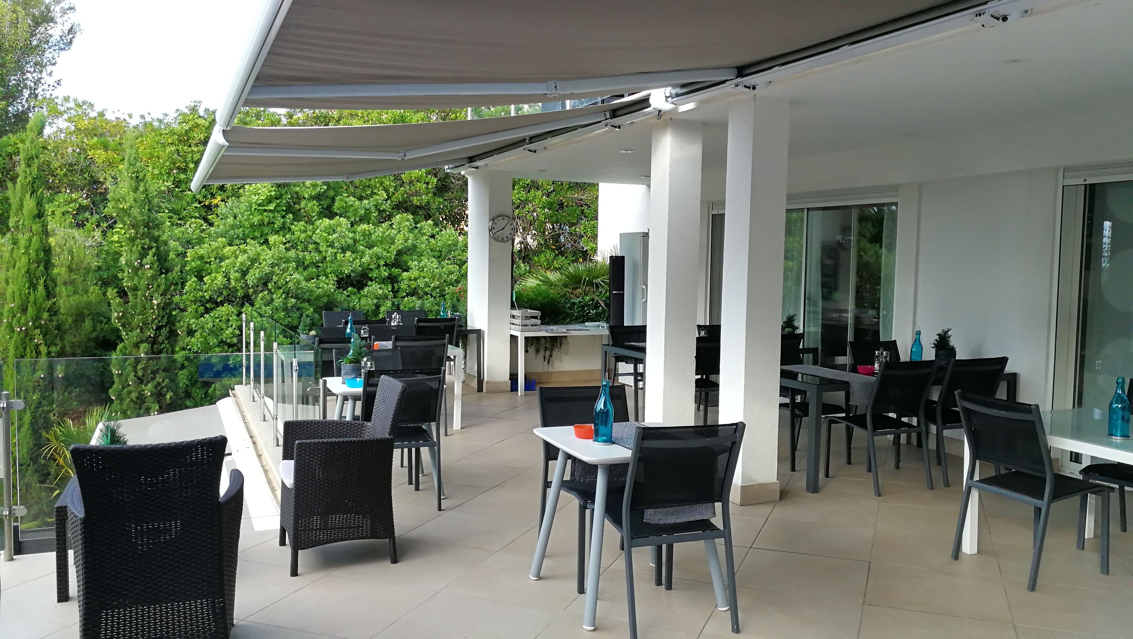 An outdoor patio area with tables and chairs, a covered seating area, and a view of greenery.
