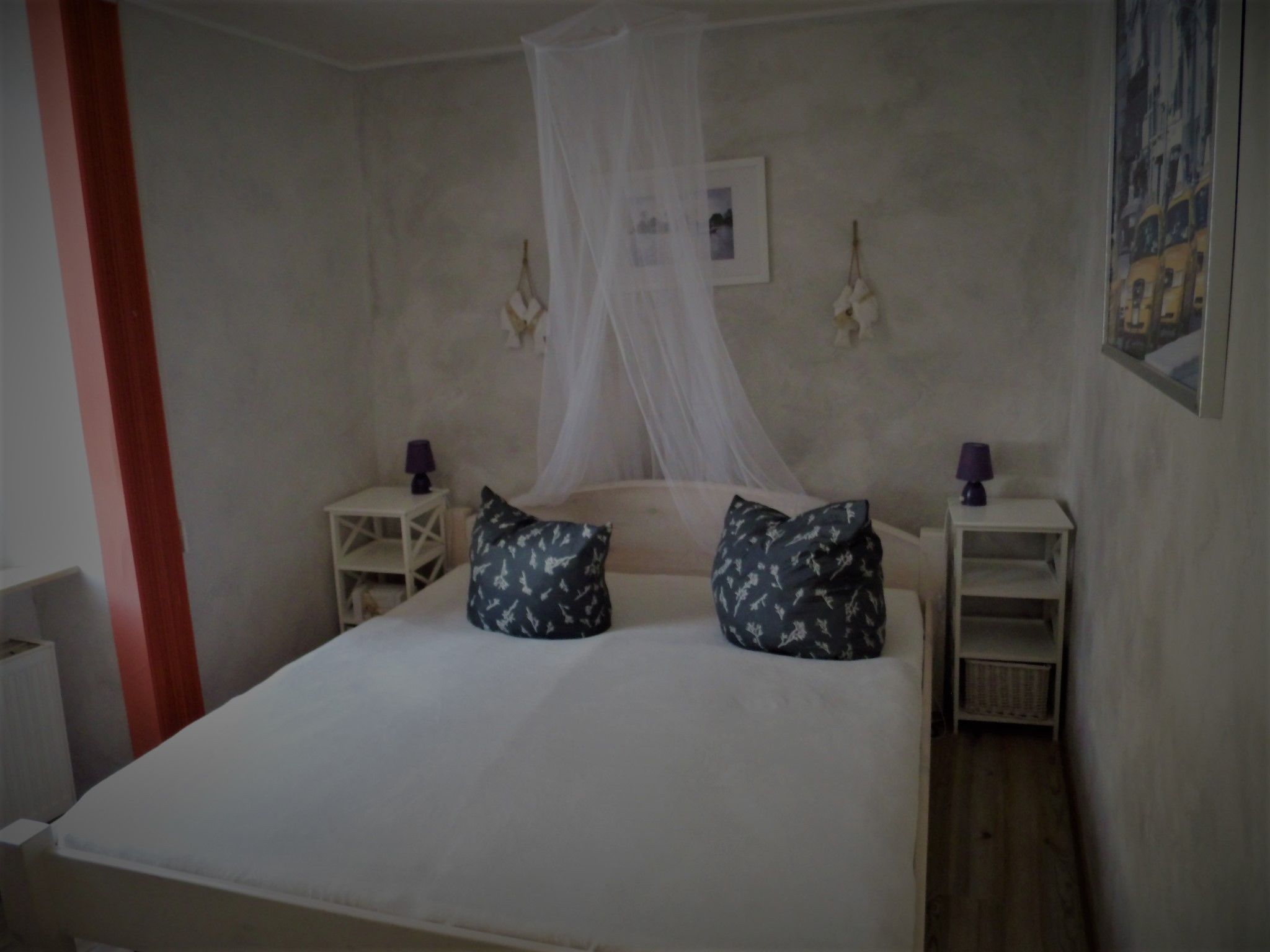 A bedroom with a bed, a nightstand, a bedside table, a lamp, a wall-mounted picture, and a window with a curtain.