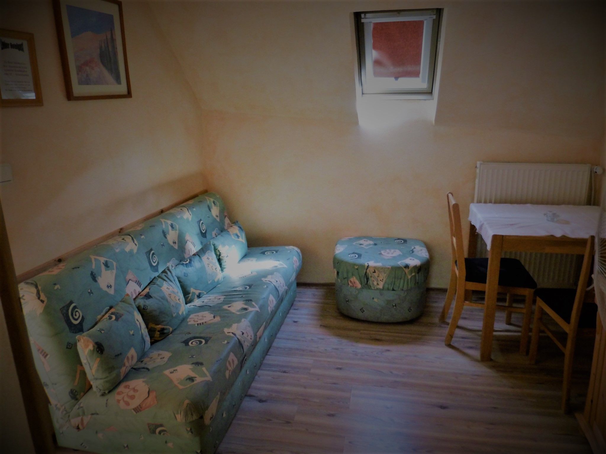 A small living room with a blue sofa, a round ottoman, a wooden chair, and a small table with a white tablecloth.