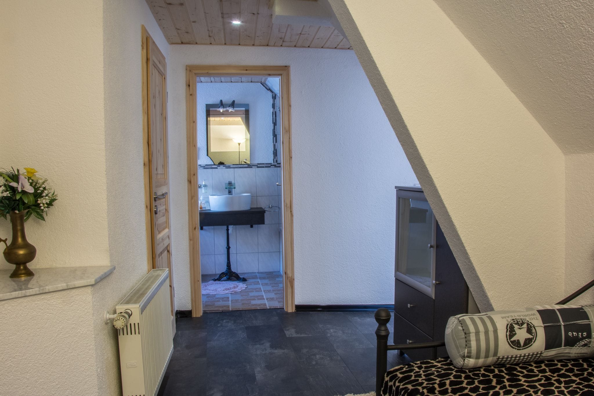 A narrow hallway leading to a bathroom with a sink and mirror. There is a radiator on the left and a bed with a patterned pillow on the right.