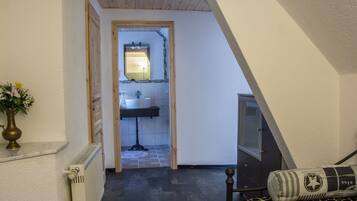 A narrow hallway leading to a bathroom with a sink and mirror. There is a radiator on the left and a bed with a patterned pillow on the right.