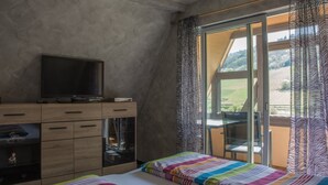 A bedroom with a bed, a TV on a wooden cabinet, and a balcony with patterned curtains. - Zum Valwiger Herrenberg (Valwig)