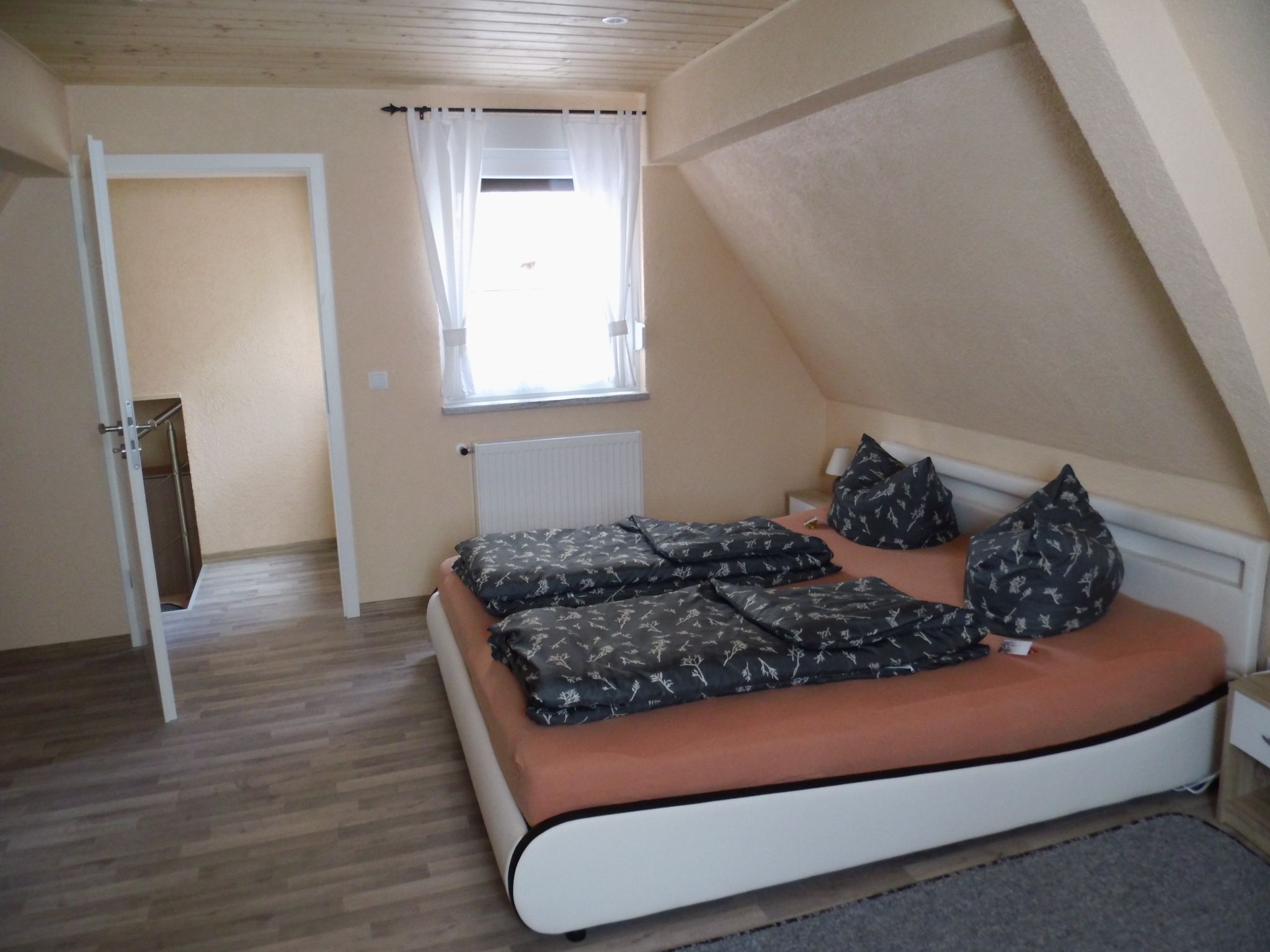 Ein Schlafzimmer mit schräg steilendem Dach, einem Bett mit gemusterten Betttextilien, einem Fenster mit Vorhängen und einer Heizung.