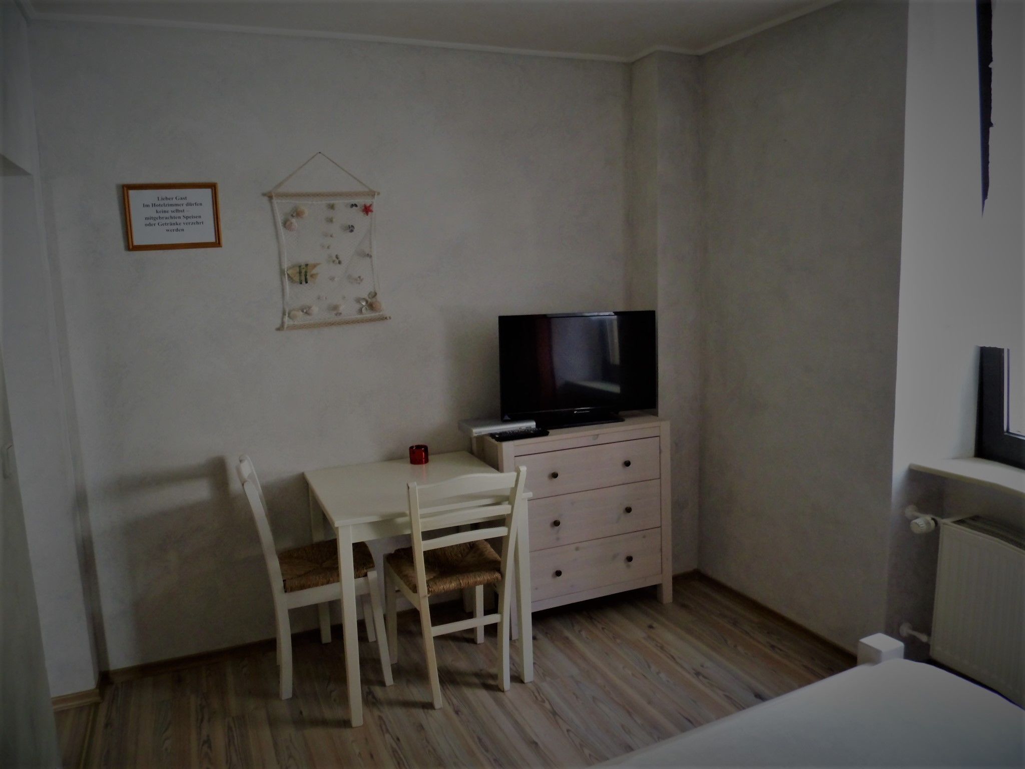 A small room with a wooden floor, a white desk with a chair, a television on a dresser, and a framed sign on the wall.