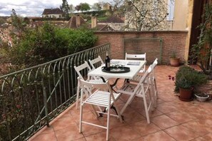 Un patio con mesa y sillas, rodeado por una barandilla metálica, con plantas en macetas y vistas al vecindario.
