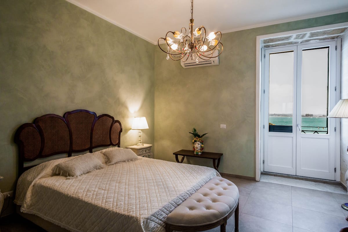 Ein Schlafzimmer mit Bett, einer Chandelier, einem Nachttisch und Blick ins Freie.