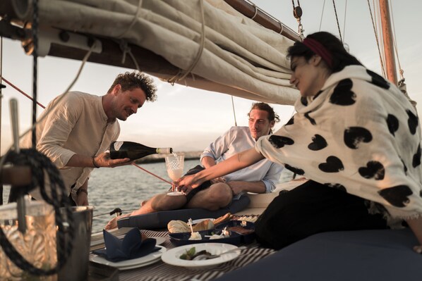 A group of people on a boat with a table set for a meal, a bottle of champagne, and a view of the water.