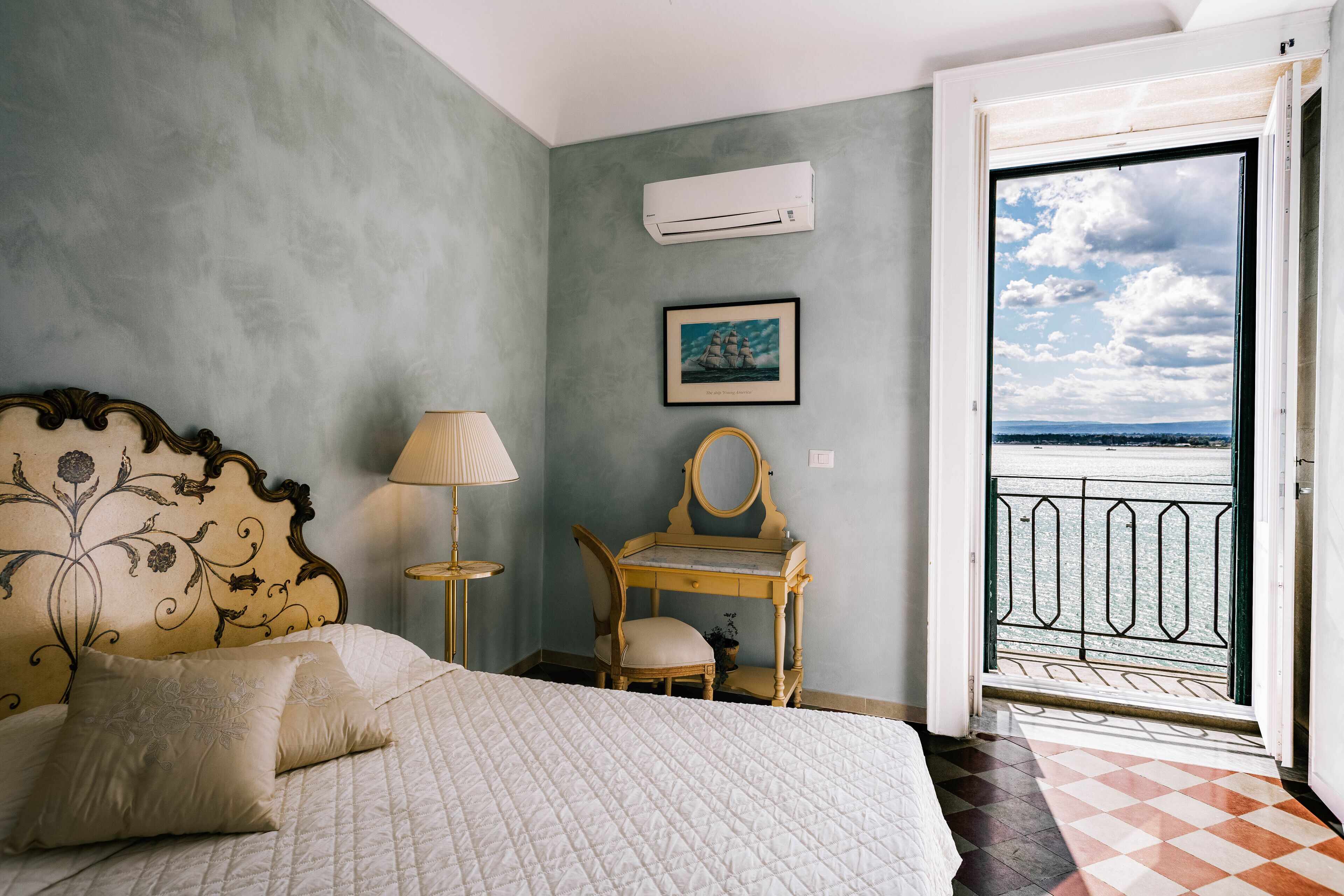 A bedroom with a bed, a desk, a chair, a lamp, a picture, and a view of the outside.