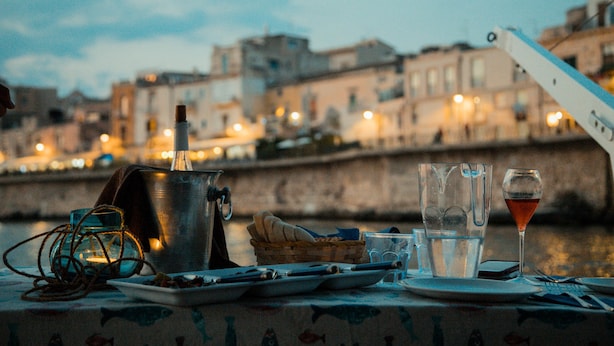 A table set with a bottle, glasses, a basket, and a plate, overlooking a waterfront with buildings in the background. - Dimora di Ulisse (Syracuse)