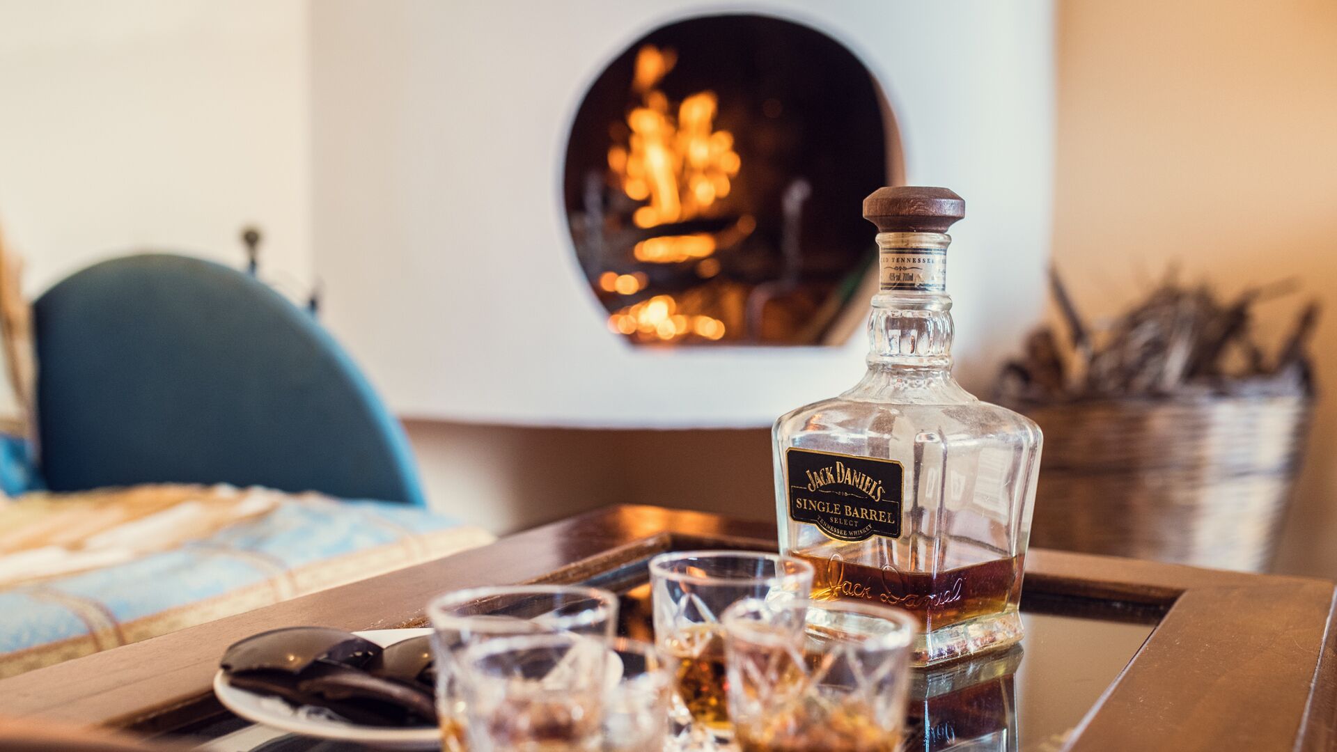 Una botella de whiskey Jack Daniel's sobre una bandeja con vasos, una silla azul y una chimenea al fondo.