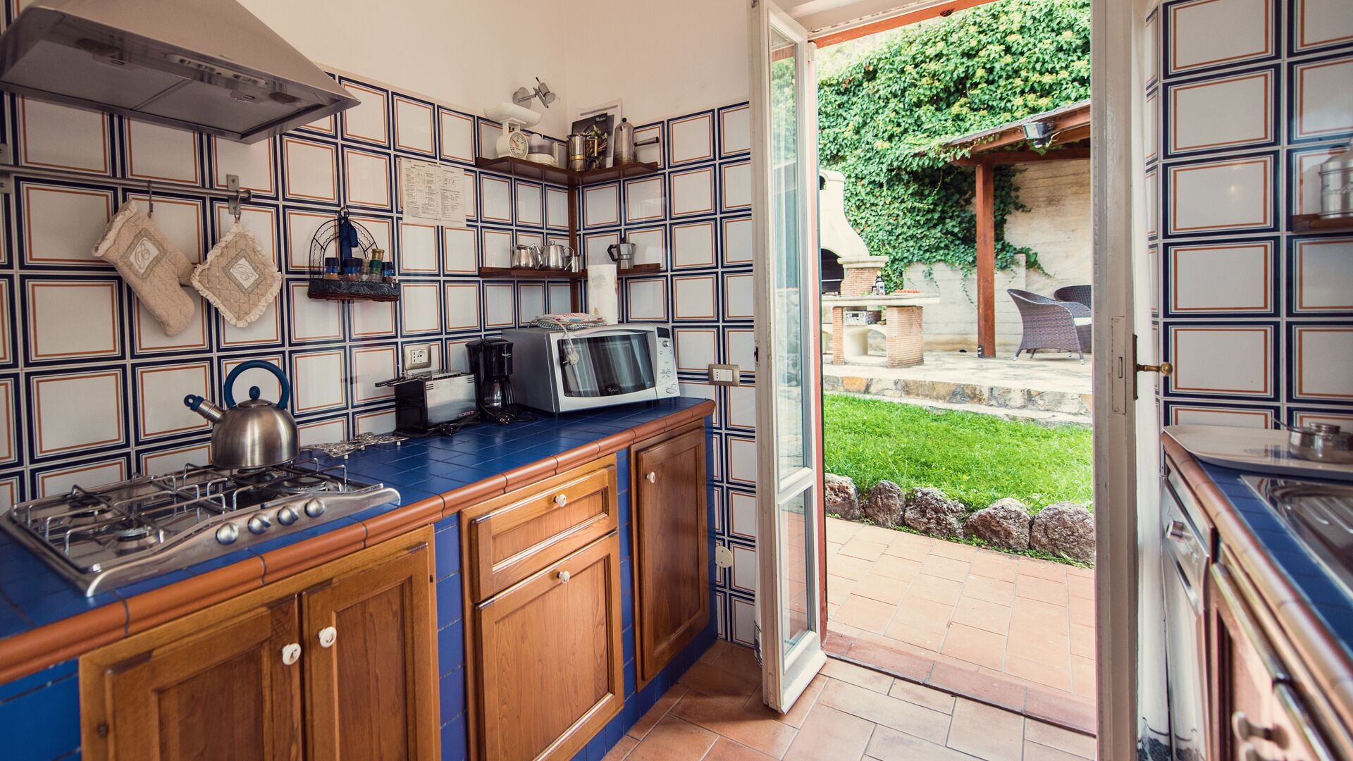 Een keuken met een blauw aanrecht, houten kastjes en een gasfornuis. Op het aanrecht staat een magnetron en een waterkoker. De keuken heeft een betegelde spatwand met een geometrisch patroon.