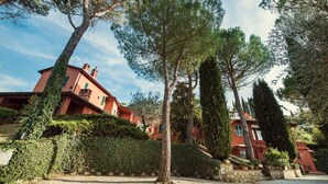 Een villa in mediterrane stijl met een rode gevel, omringd door weelderige begroeiing en hoge cipressen.