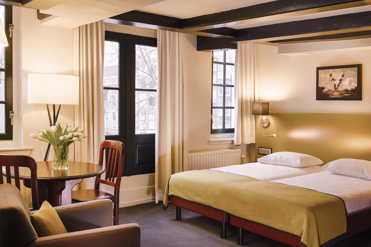 Una habitación de hotel con una cama, una silla, una mesa, una lámpara y un ventanal con cortinas.