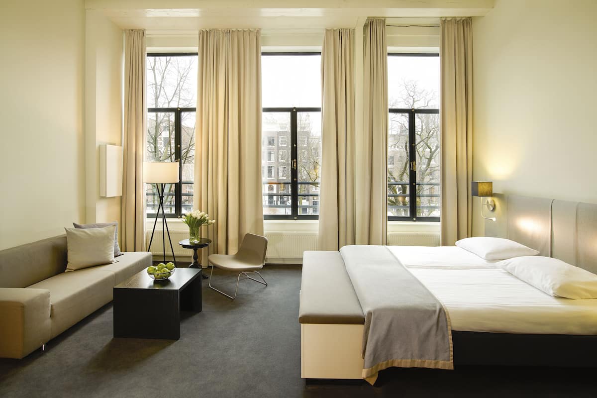 Habitación de hotel con cama, sofá, mesa de centro y una silla. Se observan ventanas con cortinas, una lámpara y vistas de edificios.