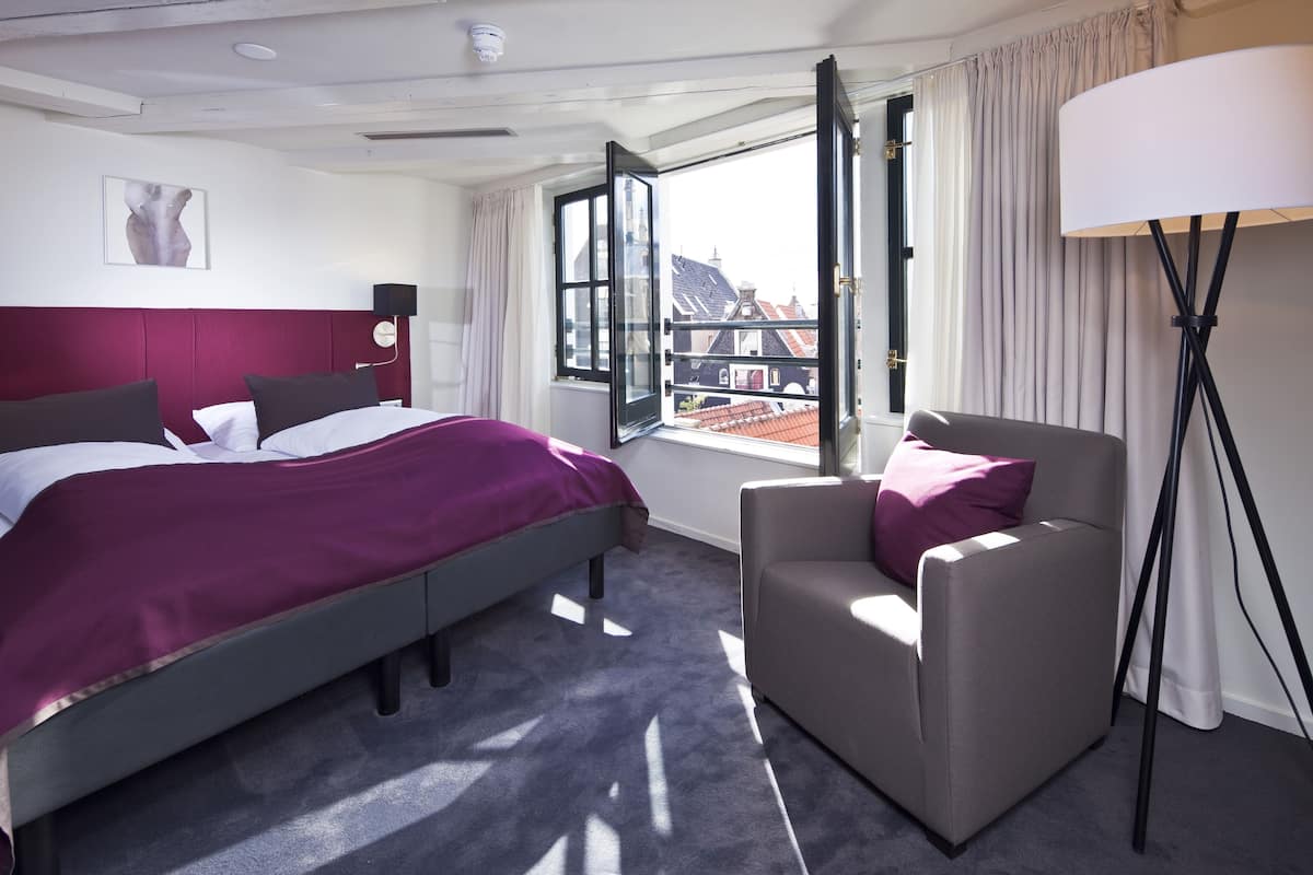 Habitación de hotel con cama, una silla, lámpara y ventana con vista a edificios.